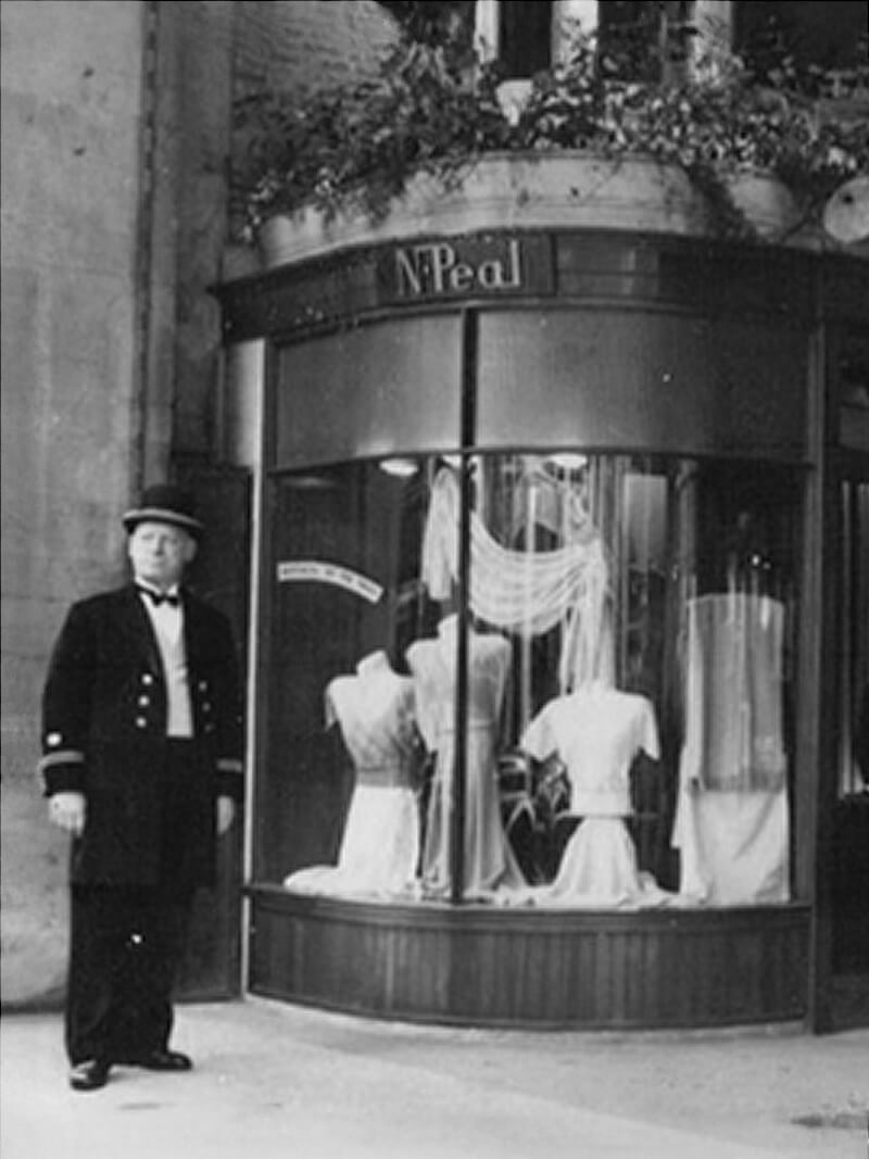 Black-and-white photograph of a doorman standing outside an N. Peal shop, with a curved display window showcasing elegant garments on mannequins and decorative drapery inside the storefront