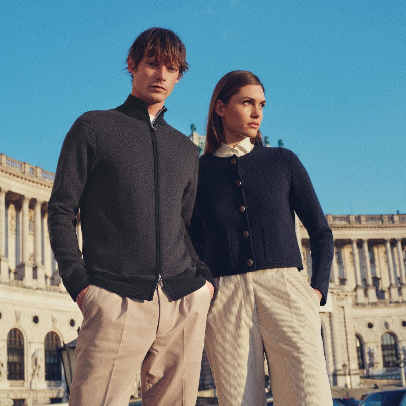 Two people standing in front of a classical building with a clear blue sky wearing N.Peal cashmere.