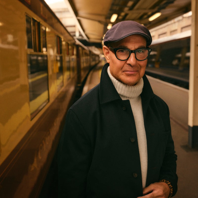 Stanley Tucci standing on a train platform beside a vintage carriage, wearing a flat cap, glasses, a dark jacket, and a cream turtleneck sweater.
