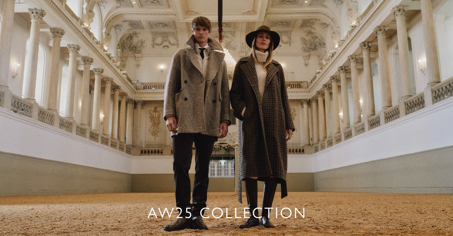 Man and woman standing side by side in an ornate grand hall with high decorated ceilings and classical columns, both wearing long textured coats; the man in a double-breasted grey coat and tie, the woman in a houndstooth coat with a turtleneck sweater and wide-brim hat.