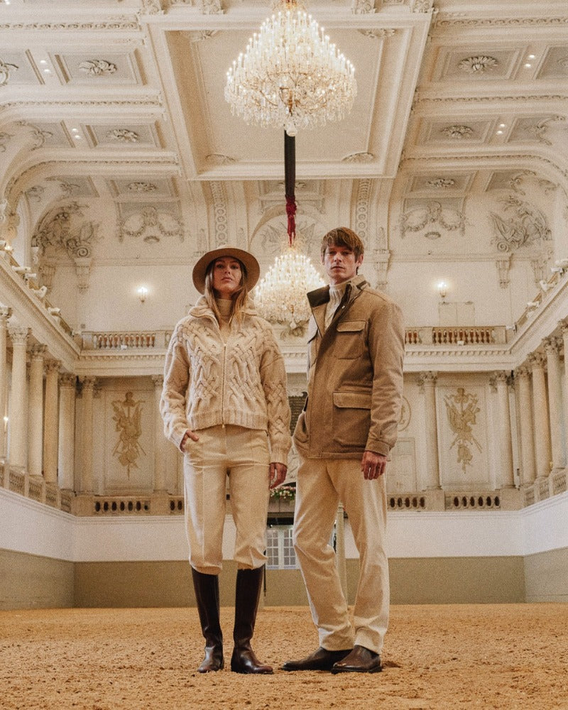 Man and woman standing in an ornate grand hall beneath chandeliers, both dressed in neutral-toned outfits; the woman in a cable-knit cardigan, cream trousers, tall riding boots, and a wide-brim hat, and the man in a beige jacket layered over a shirt and tie with matching trousers and boots