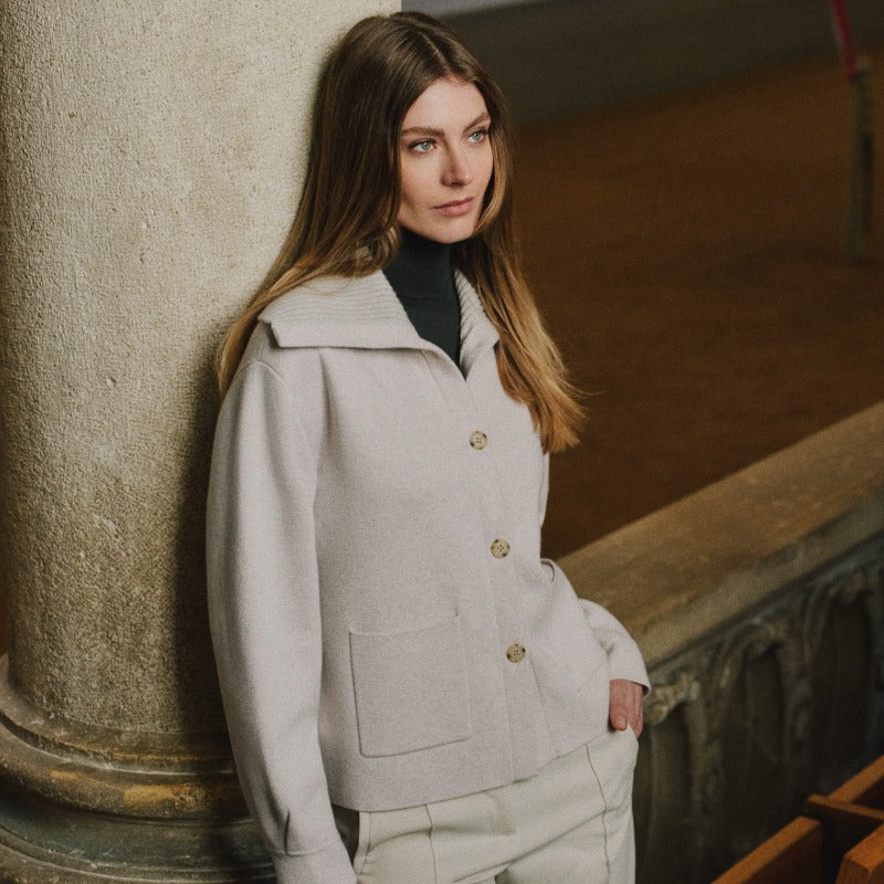 Woman leaning against a stone column wearing a light beige buttoned jacket with large front pockets, styled over a black turtleneck and cream trousers