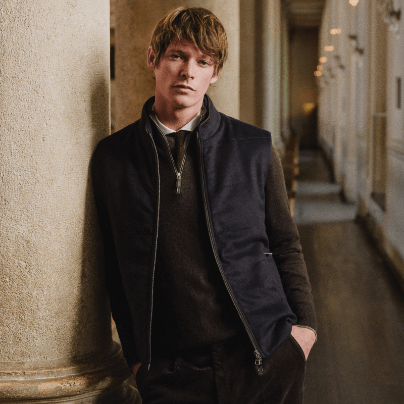 Man leaning against a stone column in a grand hallway, wearing a dark zip-up gilet layered over a sweater, shirt, and tie, with hands in his pockets