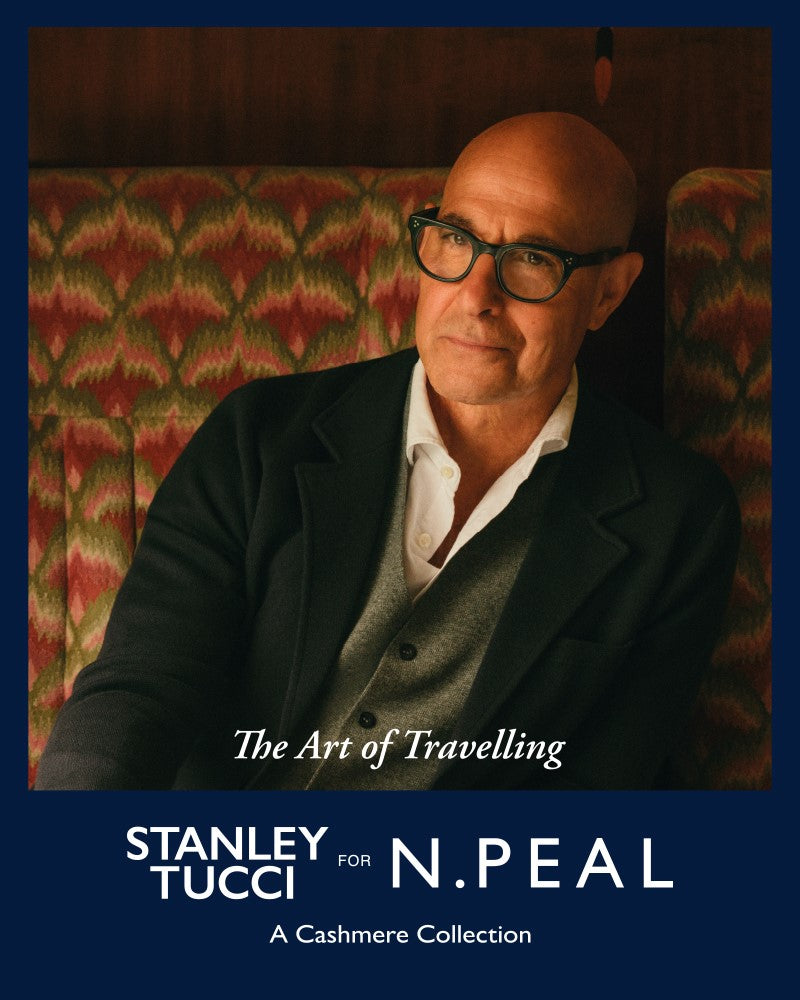 Stanley Tucci wearing glasses, a blazer, and a cashmere knit, seated against a patterned background with text overlay reading “The Art of Travelling – Stanley Tucci for N.Peal: A Cashmere Collection.”
